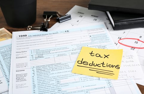 A yellow sticky note with the words “tax deductions” underlined on it. The sticky note is attached to a 1040 tax form, which is on top of a calendar with April 15 circled in red.