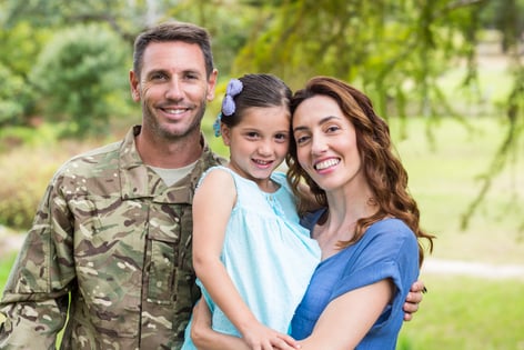 A military veteran in fatigues poses with their family in a park, with green trees in the background.