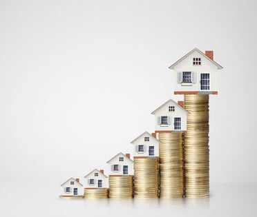 Close-up of six tiny model houses, each resting on a stack of gold coins. The stacks of coins are progressively taller from left to right, elevating their respective houses higher.