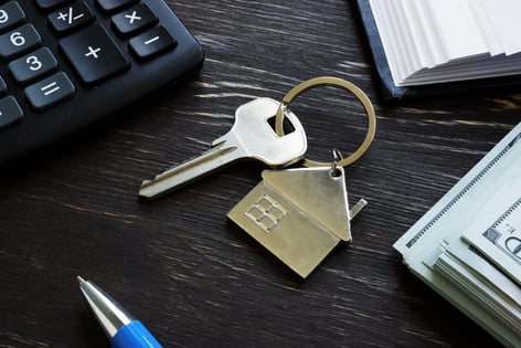 A key and a metal cutout of a house on a keyring atop the black woodgrain surface of a desk, where parts of a keyboard, a book, and a stack of cash can be seen.