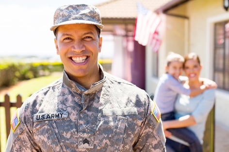 A smiling person wearing a U.S. Army uniform stands outside of a house with their family behind them. An American flag is hanging from the side of the house.