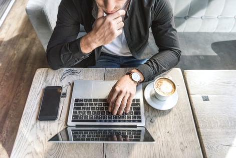 Overhead view of an individual seated at a wooden table, working on a laptop with a smartphone to one side and a latte in a white mug to the other side.